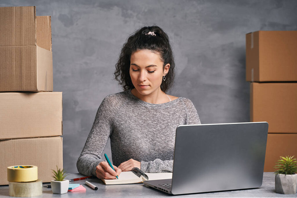 Business nainen työskentelee kotoa kannettavan tietokoneen ja muistilehtiö, valmistelee laatikot tuotteita lähettää - Valokuva, kuva
