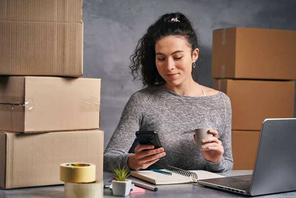 Donna d'affari che lavora da casa con laptop e smartphone, preparando scatole con prodotti da inviare - Foto, immagini