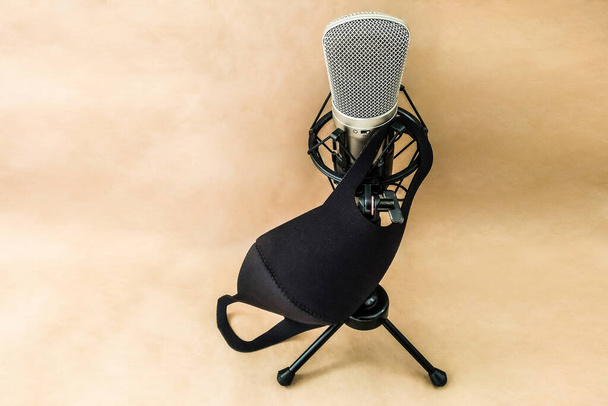 A sound recording microphone stands on a stand with a protective black mask from a coronavirus on a beige paper background with space for text. Microphone and mask from COVID-19. - Photo, Image