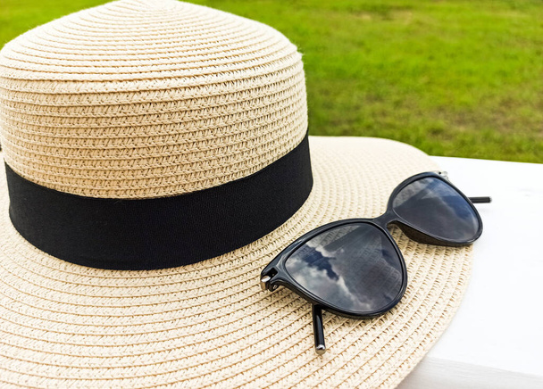 A straw female hat with sun glasses lies on a white table. Hat with a black ribbon and black women's glasses on a background of green grass. Holidays on the coast. - Photo, Image
