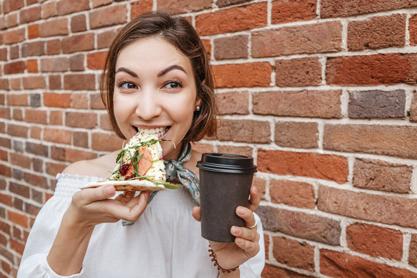 Asian girl enjoys and eats a piece of delicious hot Italian pizza against a vintage brick wall. Drinking beverage from a takeaway cup. Diet and gluttony concept - Foto, Imagen