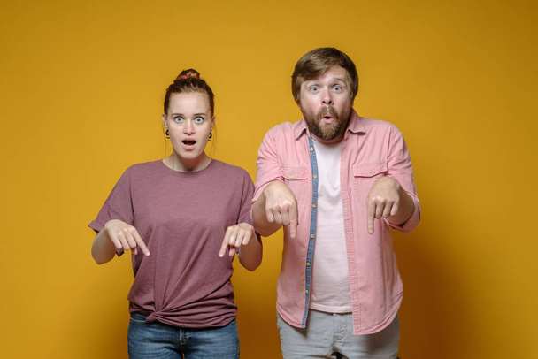 Surprised woman and man point down with their index fingers and stare wide-eyed at the camera, on a yellow background. - Photo, Image