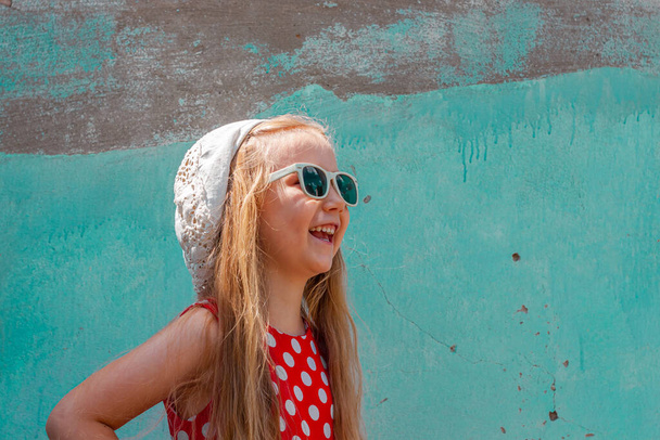 Hermosa chica feliz en un vestido de lunares rojo posando y sonriendo en la pared de hormigón turquesa background.Cute niño alegre con el pelo largo y rubio en gafas de sol y sombrero blanco al aire libre.Little modelo riendo - Foto, Imagen