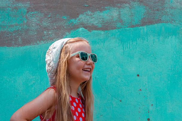 Beautiful happy girl in a red polka dot dress posing and smiling on turquoise concrete wall background.Cute joyful child with long blonde hair in sunglasses and white hat outdoor.Little model laughing - Fotoğraf, Görsel