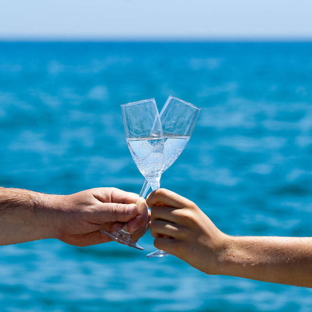 Male and female hands holding two glasses of champagne in front of blue sea and sky - Foto, afbeelding