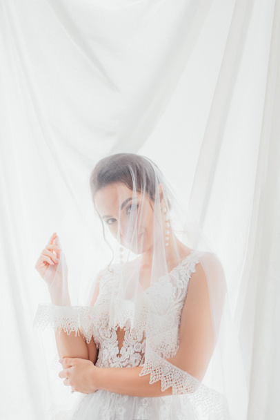 Panna młoda w sukni ślubnej i welonie patrząc na aparat w pobliżu białej tkaniny  - Zdjęcie, obraz