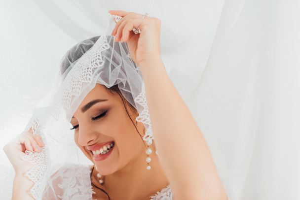 Concentration sélective de la mariée en boucles d'oreilles perle tenant voile de dentelle près de tissu blanc  - Photo, image