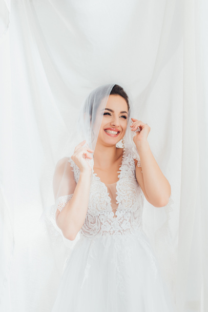 Bruneta nevěsta ve svatebních šatech a závoj dívá pryč v blízkosti bílé látky  - Fotografie, Obrázek