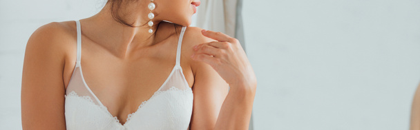 Panoramic shot of sexy bride in white bra and pearl earring touching shoulder at home - Foto, Imagem