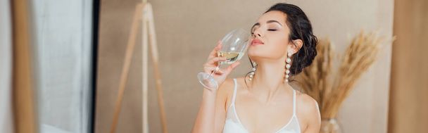 Panoramic shot of bride in bra and pearl earrings holding glass of wine at home  - Photo, Image
