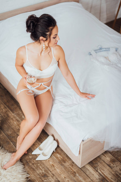 Vue aérienne de mariée en lingerie tenant un verre de vin et touchant robe de mariée sur le lit  - Photo, image