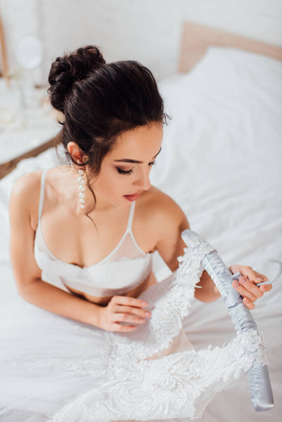 Overhead view of bride in bra and pearl earring holding hanger with wedding dress on bed  - Fotó, kép