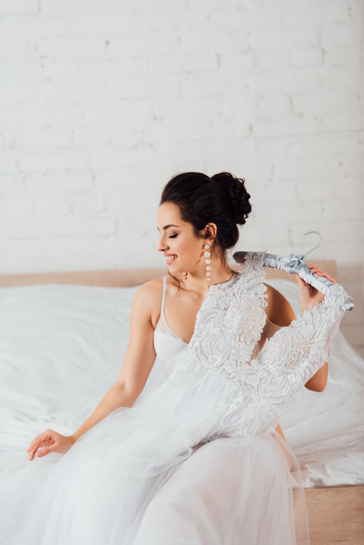 Mariée en soutien-gorge et boucles d'oreilles perle tenant cintre avec robe de mariée blanche sur le lit à la maison  - Photo, image