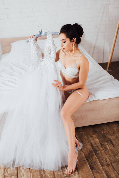 Brunette bride in lace bra touching wedding dress on hanger in bedroom - Фото, изображение