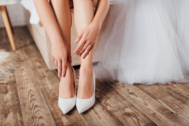 Cropped view of bride touching legs in wedding heels near dress on bed at home  - Foto, immagini