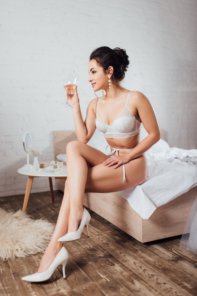 Bride in bra and shoes holding glass of wine and looking away on bed  - Foto, imagen