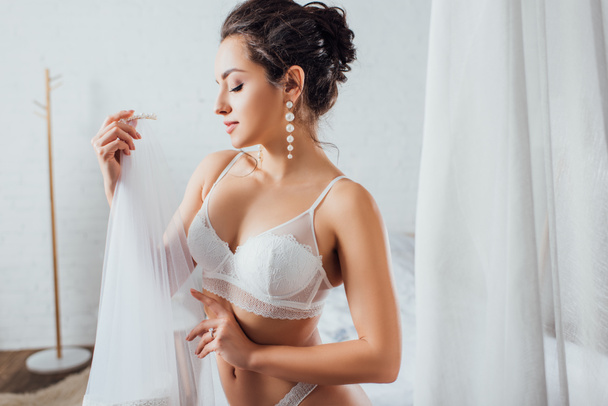 Brunette bride in lingerie holding veil at home  - Photo, Image