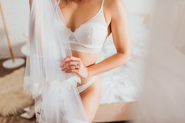 Cropped view of bride in white underwear holding lace veil in bedroom - Fotó, kép