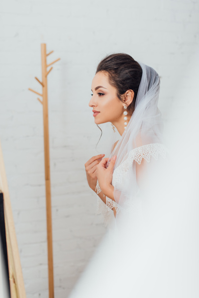Επιλεκτική εστίαση της μελαχρινής νύφης με μαργαριταρένιο σκουλαρίκι φορώντας πέπλο στο σπίτι  - Φωτογραφία, εικόνα