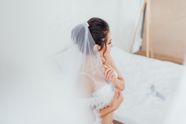 Selektiver Fokus der jungen Braut in Spitzen-BH berührt Schleier im Schlafzimmer zu Hause  - Foto, Bild