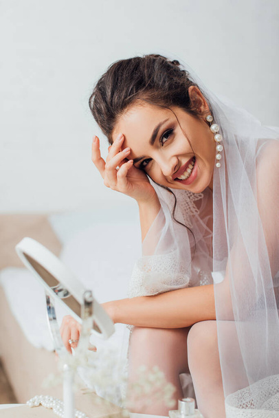 Selective focus of bride in veil looking at camera near mirror in bedroom  - Фото, изображение