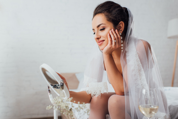 Szelektív fókusz menyasszony fátyolban nézi tükör közelében üveg bor a hálószobában - Fotó, kép