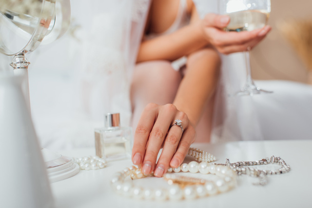 Oříznutý pohled nevěsty dotýkající se perleťového náhrdelníku a držící sklenici vína doma  - Fotografie, Obrázek
