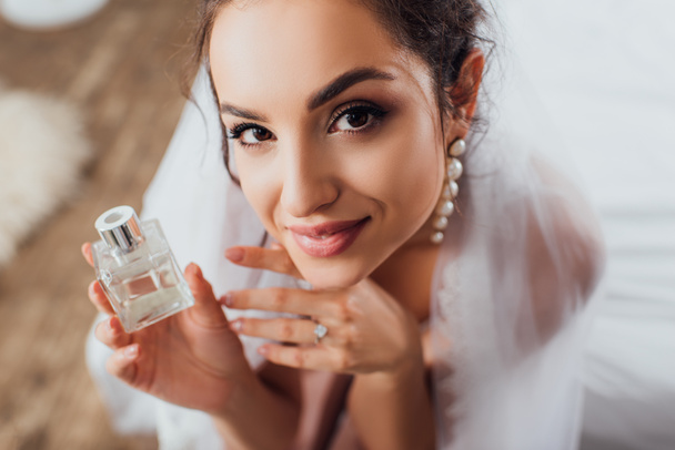 High angle view of young bride in veil looking at camera while holding bottle of perfume on bed  - Photo, Image