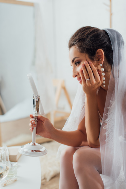 Szelektív fókusz menyasszony fülbevaló és fátyol tartó tükör közelében dohányzóasztal otthon - Fotó, kép