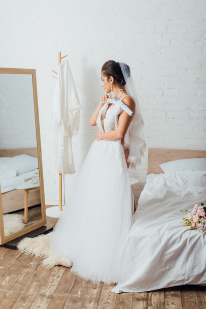 Bride in panties and veil holding hanger with wedding dress near mirror and bouquet on bed  - Zdjęcie, obraz