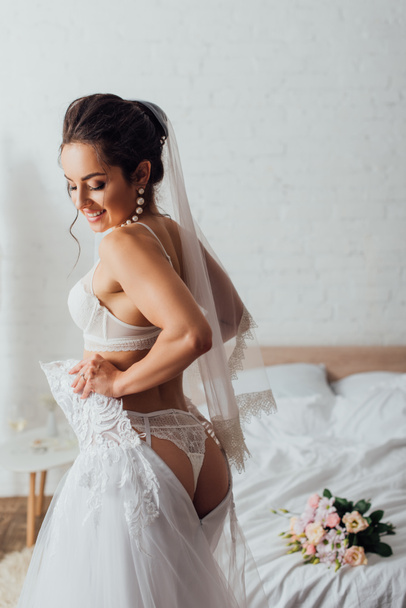 Young bride in lingerie and veil wearing wedding dress near bouquet on bed  - Photo, Image