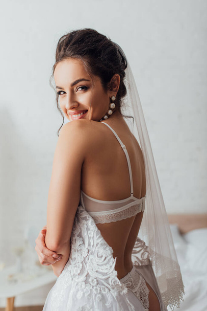 Selective focus of brunette bride in underwear, wedding dress and veil looking at camera  - Photo, Image