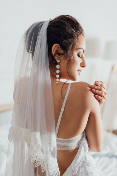 Back view of young bride in veil and wedding dress touching shoulder at home  - Foto, Imagem