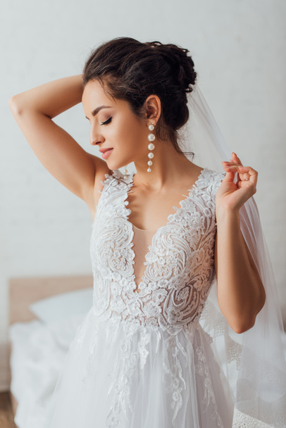 Young bride with closed eyes in lace wedding dress touching veil at home  - Photo, Image