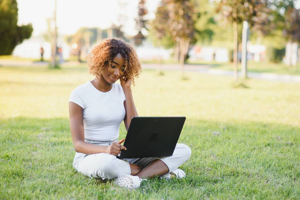 Young attractive dark-skinned college student wearing tank top and jeans sitting on the lawn at campus on sunny day, working on her thesis using laptop computer, looking busy and concentrated - Zdjęcie, obraz