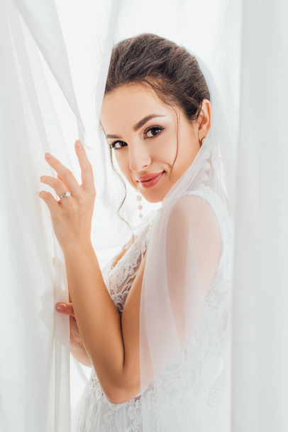 Vista lateral de la joven novia morena mirando a la cámara mientras toca las cortinas en casa  - Foto, imagen