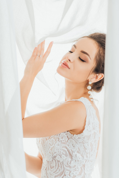 Focus selettivo della giovane sposa con gli occhi chiusi toccare tende bianche  - Foto, immagini