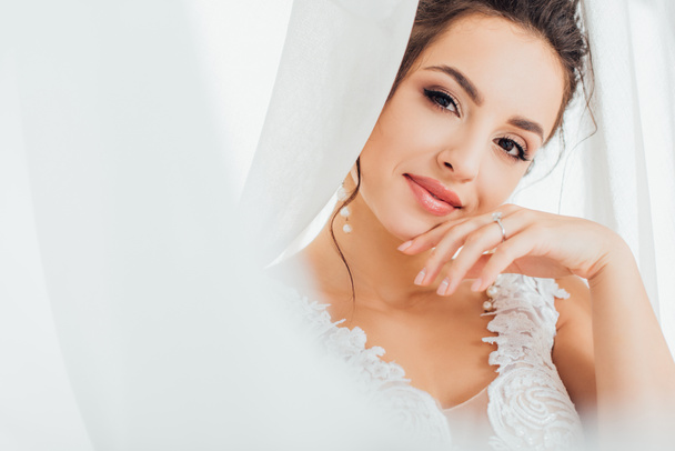 Enfoque selectivo de la novia joven en vestido de encaje mirando a la cámara cerca de cortinas blancas  - Foto, Imagen