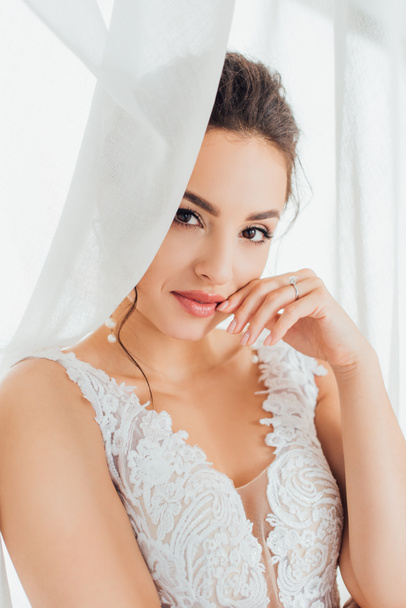 Selective focus of bride with hand near lips looking at camera near white curtains  - Photo, Image