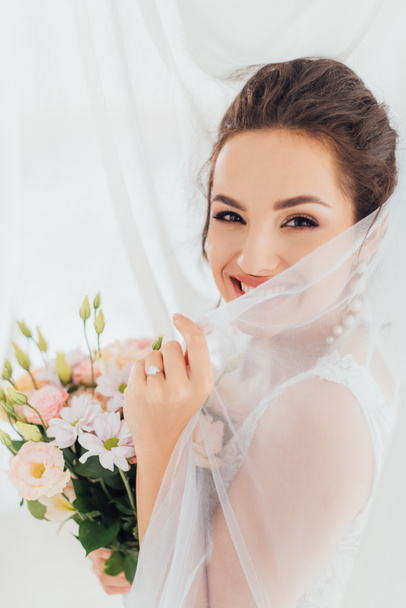 Young bride holding veil and bouquet while looking at camera near curtains  - Photo, Image