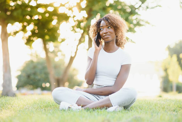 Beautiful dark skinned hipster girl talking on mobile phone sitting on green grass recreating outdoors, african american woman millennial sitting on green grass talking on cellular and good tariffs - Foto, Imagen