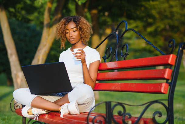 Cheerful young black girl on a wooden bench in a park is using the laptop; dazzling smiling Brazilian female outdoors on the bench with the netbook in hands and small leather bag on the left - Foto, imagen