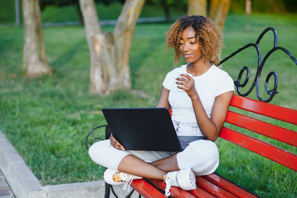 Cheerful young black girl on a wooden bench in a park is using the laptop; dazzling smiling Brazilian female outdoors on the bench with the netbook in hands and small leather bag on the left - Фото, зображення