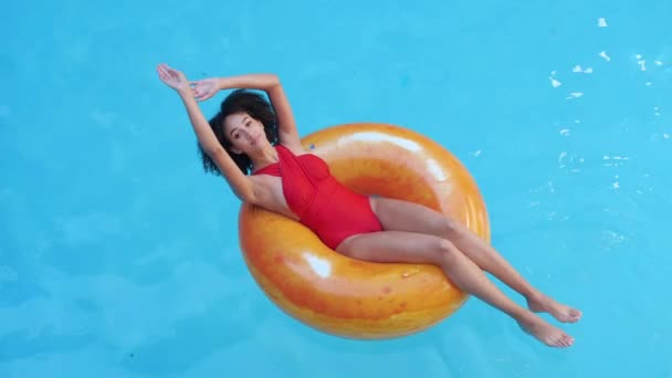 Молода задоволена чорно-волохата жінка в червоному розслаблюючому стані, піднімає руки в повітря і відчуває свободу, закриває очі, сидить в помаранчевому гумовому кільці, купається в басейні в готелі тропічний курорт за кордоном
 - Кадри, відео