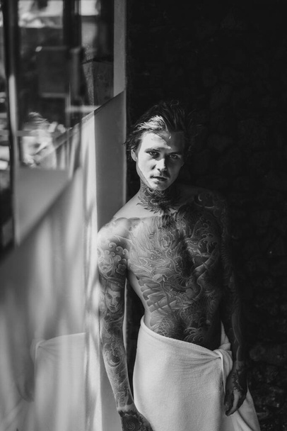 handsome young man in tattoos in the bathroom after a shower in  - Foto, afbeelding