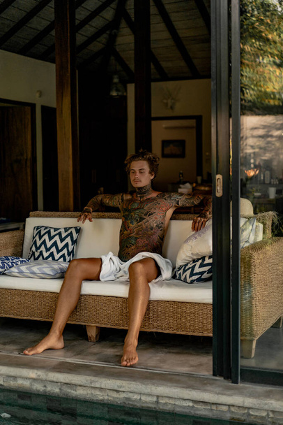 handsome young man in tattoos is resting in the living room - 写真・画像