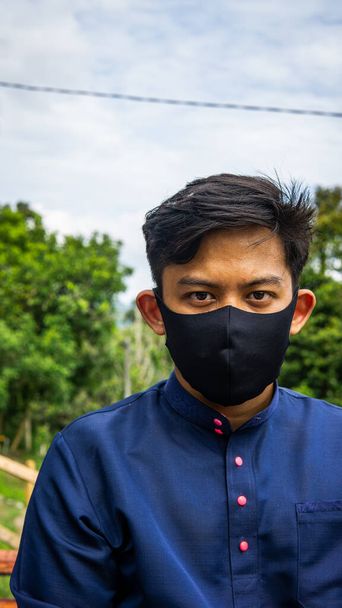 Un retrato de un joven malayo con un paño tradicional Baju Melayu con una máscara facial de tela negra con fondo verde natural. Medidas de protección para evitar la propagación de virus y enfermedades. - Foto, imagen