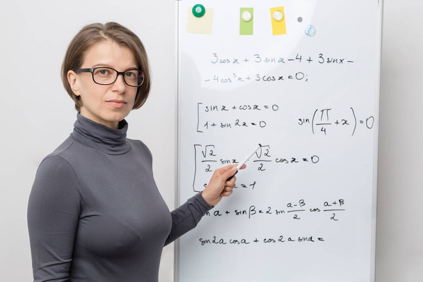 A female teacher near a white board looks at the camera and teaches online. - Foto, imagen
