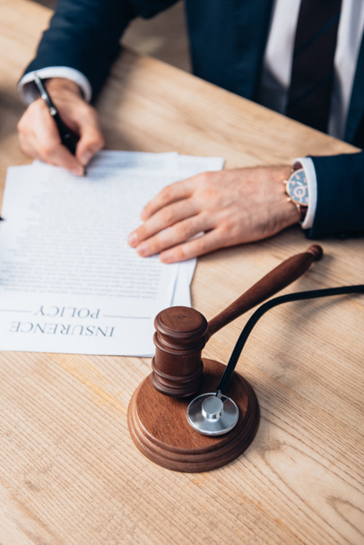 Teilansicht des Richters beim Unterschreiben von Papieren mit Versicherungsaufdruck in der Nähe von Hammer und Stethoskop auf dem Tisch - Foto, Bild