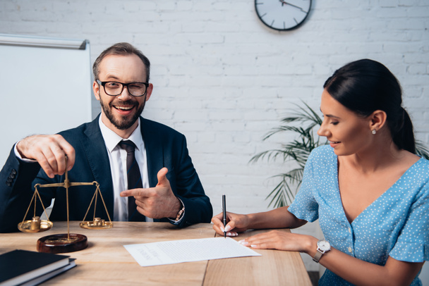 селективное внимание бородатого адвоката в очках, указывающего пальцем на весы, в то время как женщина подписывает договор страхования в офисе  - Фото, изображение